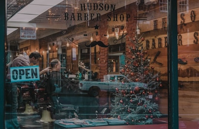 A barber shop with a festive Christmas tree inside, decorated with red ornaments. A barber is working on a customer's haircut. The shop's name and the word 'OPEN' are visible on the glass window. The interior showcases a warm, rustic decor with wooden walls and ambient lighting.