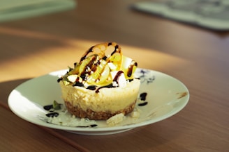 A close-up of a small cheesecake served on a white plate with a floral pattern. The cheesecake has a crumbly base and is topped with sliced fruits and a drizzle of dark syrup. The scene is lit by natural light coming from the side, casting soft shadows on the wooden table.
