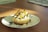 A close-up of a small cheesecake served on a white plate with a floral pattern. The cheesecake has a crumbly base and is topped with sliced fruits and a drizzle of dark syrup. The scene is lit by natural light coming from the side, casting soft shadows on the wooden table.