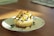 A close-up of a small cheesecake served on a white plate with a floral pattern. The cheesecake has a crumbly base and is topped with sliced fruits and a drizzle of dark syrup. The scene is lit by natural light coming from the side, casting soft shadows on the wooden table.