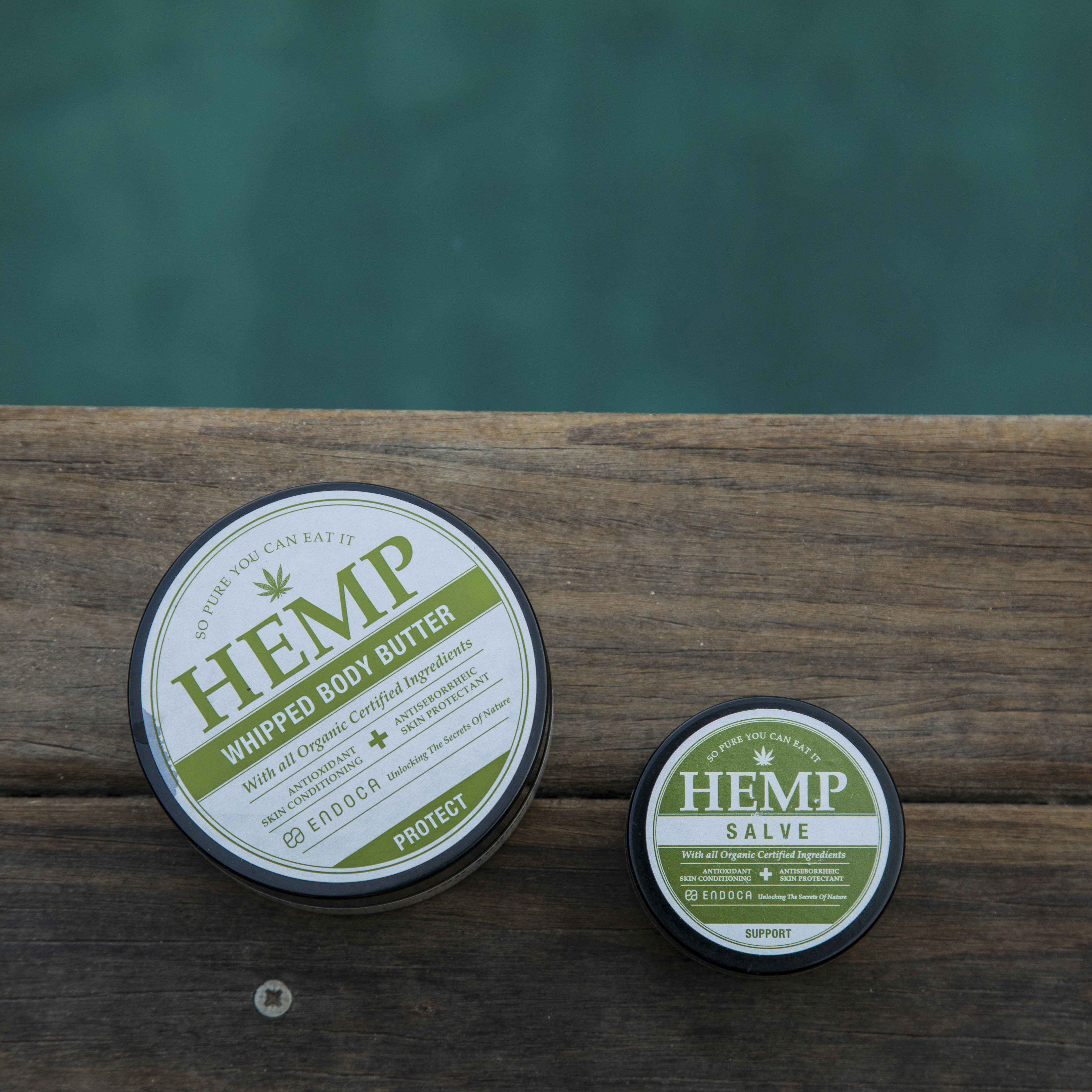 Two round containers of hemp-based skincare products placed on a wooden surface, with a calm outdoor setting. The larger container is labeled 'Hemp Whipped Body Butter' and the smaller one 'Hemp Salve'. The labels are primarily in green and white.