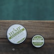 Two round containers of hemp-based skincare products placed on a wooden surface, with a calm outdoor setting. The larger container is labeled 'Hemp Whipped Body Butter' and the smaller one 'Hemp Salve'. The labels are primarily in green and white.