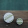 Two round containers of hemp-based skincare products placed on a wooden surface, with a calm outdoor setting. The larger container is labeled 'Hemp Whipped Body Butter' and the smaller one 'Hemp Salve'. The labels are primarily in green and white.