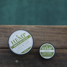 Two round containers of hemp-based skincare products placed on a wooden surface, with a calm outdoor setting. The larger container is labeled 'Hemp Whipped Body Butter' and the smaller one 'Hemp Salve'. The labels are primarily in green and white.