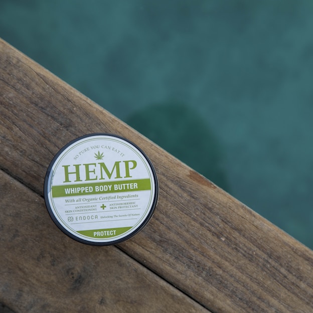 A round container of hemp whipped body butter is placed on a wooden surface. The label highlights that it contains organic certified ingredients and promotes its properties such as being an antioxidant and skin conditioner. The background features a calming, blurred teal color.