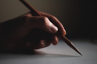 A hand is holding a sharpened pencil, poised over a light surface, suggesting an act of drawing or writing. The focus is on the careful grip of the fingers on the pencil, with dim lighting surrounding, highlighting the details of the skin and the texture of the pencil.