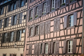 A traditional building showcases timber framing with beige and pink walls. The structure features multiple windows, most with closed shutters, and a classic streetlamp protrudes from the wall.