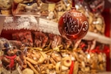 A festive holiday scene featuring a variety of cookies and confections wrapped in clear packaging and tied with ribbons. A prominent element is a red Christmas ornament inscribed with 'Joyeux Noël.' The foreground includes a chocolate snowman wrapped in plastic.