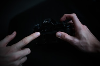 Two hands are interacting with a vintage camera in a dimly lit setting, creating a sense of focus and delicacy. The surrounding darkness emphasizes the hands and the camera, adding a dramatic mood.