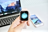 A smart watch with a pink band held in front of a laptop displaying a mountainous wallpaper. In the background, a smartphone with a similar aesthetic is visible on a white surface.
