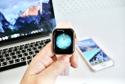 A smart watch with a pink band held in front of a laptop displaying a mountainous wallpaper. In the background, a smartphone with a similar aesthetic is visible on a white surface.