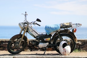 A rusty, old motorcycle is parked by a stone wall overlooking a serene ocean. The motorcycle has a makeshift seat cover made of a blue material, and a helmet rests on the ground next to it.