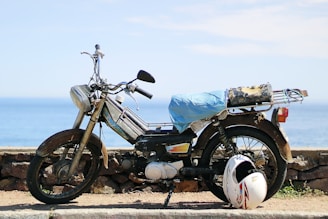 A rusty, old motorcycle is parked by a stone wall overlooking a serene ocean. The motorcycle has a makeshift seat cover made of a blue material, and a helmet rests on the ground next to it.