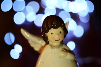 A ceramic figurine of an angel with a smiling face and brown hair is positioned against a backdrop of soft blue bokeh lights. The angel has a glossy finish and small wings visible on its back.