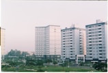 Several mid-rise apartment buildings with a uniform architectural style, featuring numerous windows and balconies, are lined up next to each other. The buildings are surrounded by small park areas with green lawns and scattered trees. The sky is overcast with a soft light, contributing to a calm atmosphere.