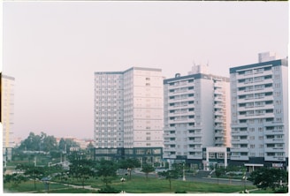 Several mid-rise apartment buildings with a uniform architectural style, featuring numerous windows and balconies, are lined up next to each other. The buildings are surrounded by small park areas with green lawns and scattered trees. The sky is overcast with a soft light, contributing to a calm atmosphere.