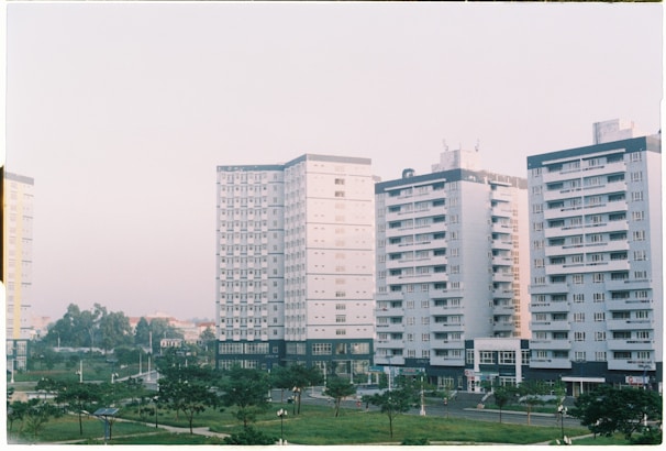 Several mid-rise apartment buildings with a uniform architectural style, featuring numerous windows and balconies, are lined up next to each other. The buildings are surrounded by small park areas with green lawns and scattered trees. The sky is overcast with a soft light, contributing to a calm atmosphere.