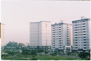 Several mid-rise apartment buildings with a uniform architectural style, featuring numerous windows and balconies, are lined up next to each other. The buildings are surrounded by small park areas with green lawns and scattered trees. The sky is overcast with a soft light, contributing to a calm atmosphere.