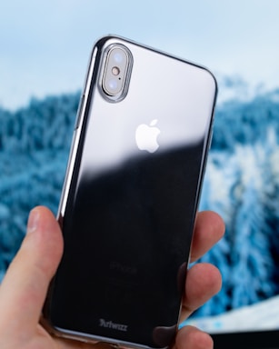 A person is holding a shiny, reflective smartphone with the Apple logo on the back. The phone has dual cameras on the upper left corner and is encased in a transparent Artwizz case. The background is blurred, showing a natural winter scene with snow-covered trees.