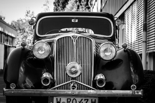 A classic vintage car with prominent round headlights and a distinct chrome grille, captured in black and white. The car is parked outdoors, with modern buildings in the background, highlighting its retro charm.