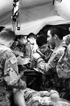 Several people in military uniforms are gathered closely together, possibly in a medical or field training environment. They appear to be engaged in a focused task, with one person writing and others observing or participating. A person’s feet can be seen in the foreground, suggesting medical attention.
