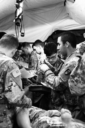 Several people in military uniforms are gathered closely together, possibly in a medical or field training environment. They appear to be engaged in a focused task, with one person writing and others observing or participating. A person&rsquo;s feet can be seen in the foreground, suggesting medical attention.