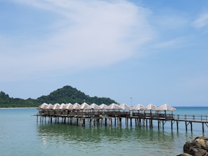 A series of wooden huts with thatched roofs extends over the calm turquoise water, connected by a pier. In the background, a lush green mountain rises against a clear blue sky, creating a serene and picturesque coastal scene.