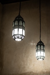 Two intricately designed hanging lanterns emit a soft, glowing light against a textured beige wall. The lanterns feature detailed metalwork with geometric and floral patterns, suspended from chains.