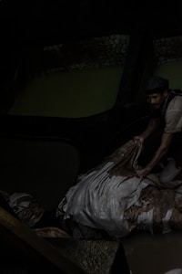 A person appears to be working in a dark, industrial environment. The individual is handling what seems to be a pile of fabric or hides, possibly in a tannery. The setting is dimly lit, with muted water and brickwork visible in the background.