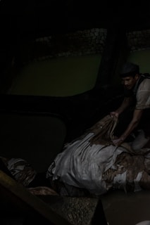 A person appears to be working in a dark, industrial environment. The individual is handling what seems to be a pile of fabric or hides, possibly in a tannery. The setting is dimly lit, with muted water and brickwork visible in the background.