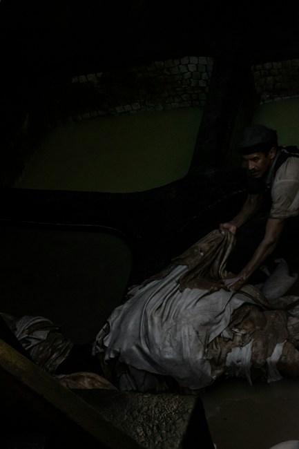 A person appears to be working in a dark, industrial environment. The individual is handling what seems to be a pile of fabric or hides, possibly in a tannery. The setting is dimly lit, with muted water and brickwork visible in the background.