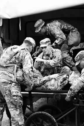 A group of soldiers in camouflage uniforms are tending to an injured comrade on a stretcher. The scene suggests a military exercise or a training environment, as the soldiers are actively engaged in care and communication.
