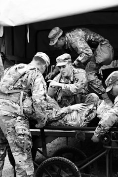 A group of soldiers in camouflage uniforms are tending to an injured comrade on a stretcher. The scene suggests a military exercise or a training environment, as the soldiers are actively engaged in care and communication.