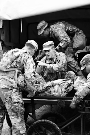 A group of soldiers in camouflage uniforms are tending to an injured comrade on a stretcher. The scene suggests a military exercise or a training environment, as the soldiers are actively engaged in care and communication.