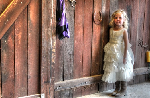 A young girl in a white, lacey dress and brown boots stands near a rustic wooden barn wall. A purple cloth hangs on a hook nearby, and a horseshoe is mounted on the wall. Ambient lighting creates a warm, nostalgic ambiance.