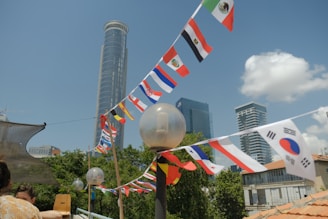 A cityscape featuring modern skyscrapers with strings of international flags strung between poles. The foreground shows assorted flags from different countries against a bright blue sky with a few clouds. A person with short hair is partially visible, standing at the side, and some foliage is visible in the background.