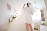 A person in a white robe and veil is standing in a bright, elegantly decorated room, lifting a white wedding dress from a hanger. The room has pastel-colored walls, a chandelier, and a window with curtains allowing soft light to filter in. A vase with pink flowers is on a dresser beside a framed picture.