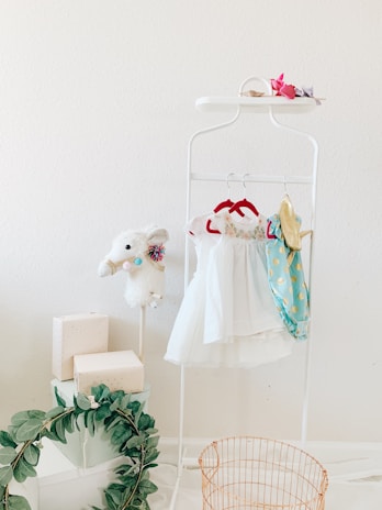 A white clothing rack holds several children's dresses, including a white dress with floral embroidery and a blue dress with gold polka dots. A plush white llama with colorful decorations is perched on the left side. There are gift boxes stacked on the floor and a decorative green wreath. A gold wire basket sits in the foreground, and the wall behind is light-colored.