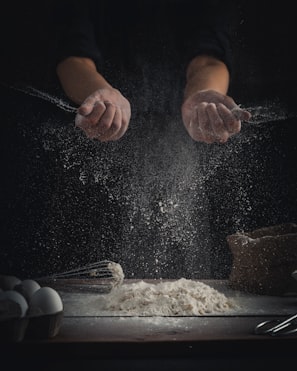 person pouring flour on table beside eggs and whisk