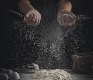 person pouring flour on table beside eggs and whisk