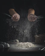 person pouring flour on table beside eggs and whisk