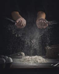 Hands are sprinkling flour over a wooden surface. A pile of flour sits on the table alongside baking tools, such as a whisk, and a carton of eggs. The scene is dimly lit, creating a dramatic contrast between the dark background and the bright flour particles scattering in the air.