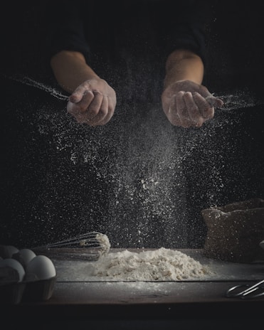 Hands are sprinkling flour over a wooden surface. A pile of flour sits on the table alongside baking tools, such as a whisk, and a carton of eggs. The scene is dimly lit, creating a dramatic contrast between the dark background and the bright flour particles scattering in the air.