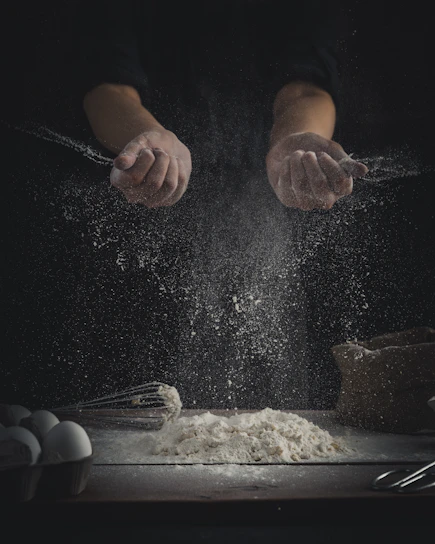 A close-up of hands dusting flour over a rustic wooden table with baking tools scattered around.