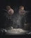 Hands are sprinkling flour over a wooden surface. A pile of flour sits on the table alongside baking tools, such as a whisk, and a carton of eggs. The scene is dimly lit, creating a dramatic contrast between the dark background and the bright flour particles scattering in the air.