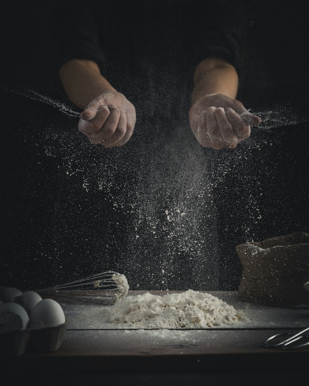 person pouring flour on table beside eggs and whisk