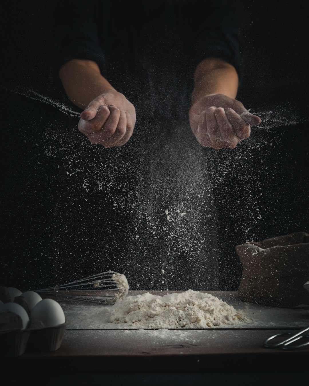 person pouring flour on table beside eggs and whisk