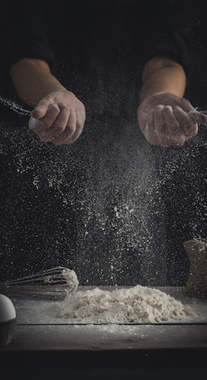 person pouring flour on table beside eggs and whisk