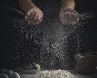 person pouring flour on table beside eggs and whisk