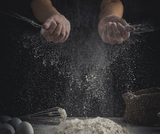 person pouring flour on table beside eggs and whisk